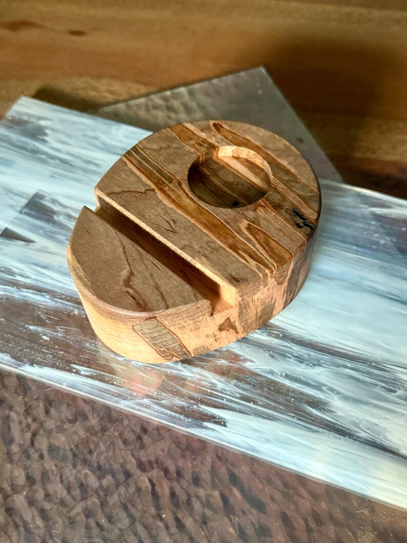 May include: A wooden phone stand with a natural wood grain pattern. The stand has a slot to hold a phone and a circular opening. The wood tones are light and dark brown. The stand is on a patterned surface.