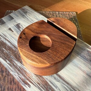 May include: A handcrafted wooden phone stand and charging station. The stand is made of dark brown wood with a circular opening and a slot for the phone. The stand is on a decorative surface.