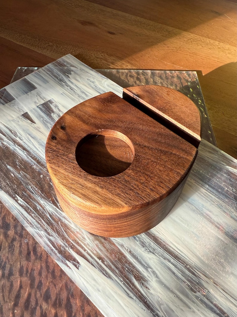 May include: A handcrafted wooden phone stand and charging station. The stand is made of dark brown wood with a circular opening and a slot for the phone. The stand is on a decorative surface.