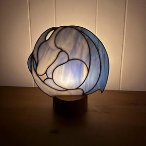 May include: A stained glass lamp with a blue and white design, set on a wooden base. The lamp has a rounded shape with a swirling pattern, and the light shines through the glass, creating a soft glow. The lamp is a decorative home decor item.