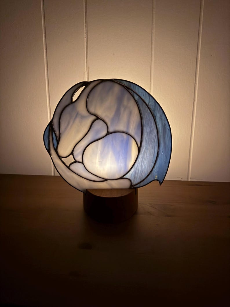 May include: A stained glass lamp with a blue and white design, set on a wooden base. The lamp has a rounded shape with a swirling pattern, and the light shines through the glass, creating a soft glow. The lamp is a decorative home decor item.