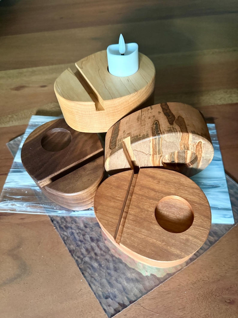 May include: A collection of wooden candle holders in various shapes and wood grains. One holder has a slot for a phone and a small, white LED candle. The other holders have circular cutouts for candles. The wood tones range from light to dark.