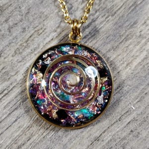 May include: A gold-toned pendant necklace with a circular, resin-filled pendant. The pendant features a gold spiral design over a mix of colorful glitter and iridescent flakes. The necklace chain is also gold.