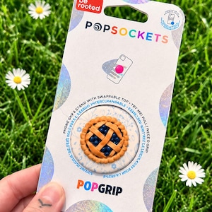 May include: A PopSockets PopGrip phone accessory in its original white packaging. The swappable top features a three-dimensional miniature blueberry pie design with a golden-brown lattice crust and dark blue berries on a light blue floral background.
