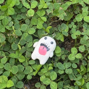 May include: A white, textured, ghost-shaped charm with two small black eyes, a wide open mouth featuring black gums and white teeth, and a large, protruding pink tongue. A small silver metal loop is attached to the top of the ghost figure.