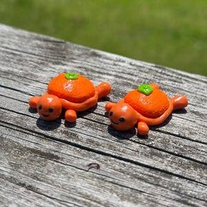 May include: Two small turtle figurines made of orange clay. Each turtle has a textured orange shell resembling a citrus fruit, topped with a small green leaf detail. The turtles have smooth orange limbs, heads with black dot eyes, and small smiles.