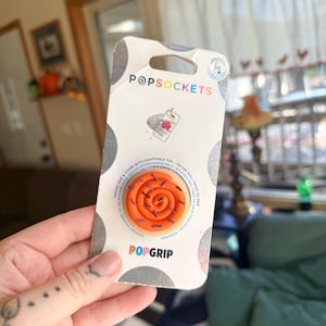 May include: A PopSockets PopGrip phone stand in retail packaging. The product is a round, orange, swirled cinnamon roll design with purple sprinkles. The white cardboard backing features the PopSockets logo and text, including instructions to pull twice to grip.