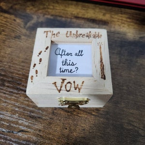 May include: A wooden box with a hinged lid and a small picture frame inside. The frame has a white background with the text "After all this time?" written in black. The box is decorated with a wand, footprints, and the words "The Unbreakable Vow".