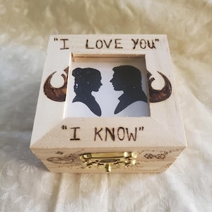May include: A wooden box with a hinged lid, featuring a framed silhouette of two figures. The box is inscribed with the phrases "I LOVE YOU" and "I KNOW". The box has a gold-colored clasp and decorative accents.