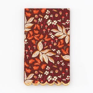 May include: A set of brown paper napkins with a gold scalloped edge. The napkins feature a repeating pattern of orange and white leaves, red mushrooms, and white flowers on a dark brown background.