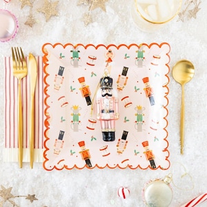 May include: A pink and white paper plate with a scalloped edge features a repeating pattern of nutcracker ornaments, mice, and drums. A pink nutcracker ornament is in the center of the plate.
