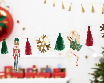 May include: A festive Christmas garland featuring a nutcracker, ballerina, gold snowflakes, and green and red tassels. The garland is strung on a gold string, with a red ornament and Christmas tree in the background.