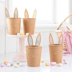 May include: Four brown paper cups with bunny ears. The ears are made of paper and have a floral pattern. The cups have a bunny face drawn on them. There are pastel colored candy sprinkles scattered around the cups.