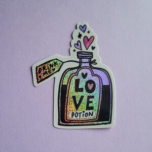 May include: A holographic sticker of a glass bottle with a black label that says "LOVE POTION" and a tag that says "DRINK ME".  Pink, red, and purple hearts are floating above the bottle.