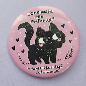 May include: A pink button featuring a black cat illustration with its tongue out. The button has white text that reads "Je ne porte pas malheur" and "ta vie était déjà de la marde". Small hearts and flowers surround the cat.