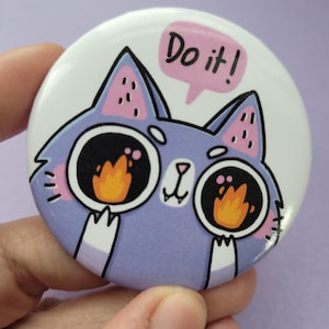 May include: A circular badge featuring a cartoon cat design. The cat is depicted in purple with large eyes and a speech bubble that reads "Do it!". The cat's eyes contain orange flames.