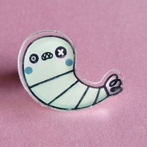 May include: A green and white cartoon ghost with a black eye patch and a black X for an eye. The ghost is wearing a white bandage and has a small arm with two circles on it.