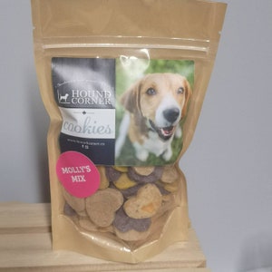Peut inclure: Un sac en papier brun avec une fenêtre en plastique transparent montrant une variété de friandises pour chiens. Le sac porte une étiquette qui dit "Hound Corner Cookies" et "Molly's Mix". L'étiquette présente également une photo d'un chien.