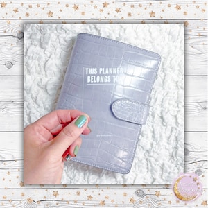 May include: A gray faux leather planner with a clear sticker that says "This Planner Belongs To".