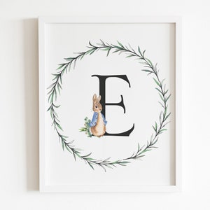 May include: A white framed print with a green leafy wreath surrounding the letter "E" in black. A small illustration of Peter Rabbit is to the left of the letter.