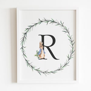 Pode incluir: Uma impressão emoldurada branca com uma ilustração em aquarela do Peter Rabbit em pé na frente da letra "R" cercada por uma coroa de folhas verdes.