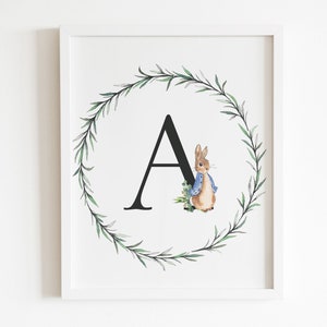 May include: A framed print with a black letter A surrounded by a green leafy wreath. A brown rabbit with a blue jacket is peeking out from behind the letter.
