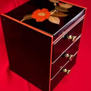 Può includere: Un mobiletto in lacca nera a tre cassetti, con bordi rossi e un motivo floreale sulla parte superiore. I cassetti sono dotati di piccoli pomelli rotondi color oro metallizzato. Il mobiletto è rettangolare con una finitura liscia e lucida.