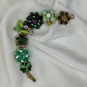 May include: A charm bracelet featuring a variety of green-toned components, including faceted glass beads, round beads in shades of lime and emerald, and ornate gold-tone metal settings with dark green crystals, finished with a gold-tone metal clasp.
