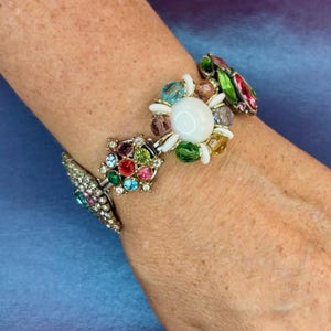 May include: A multi-colored charm bracelet featuring a central white cabochon surrounded by small faceted crystals in shades of blue, green, pink, and yellow, connected to additional decorative links set with various colorful rhinestones.