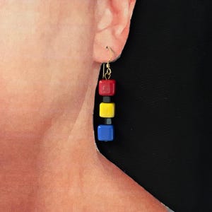 May include: A single earring with a gold hook features a dangling design of three colorful square beads. The beads are red, yellow, and blue, separated by small black beads. The earring is shown against a black background.