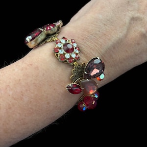 May include: A gold tone bracelet featuring a cluster of red, pink and purple rhinestones. The bracelet is made of metal and has a delicate, feminine design.