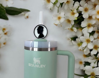 May include: A green Stanley travel mug with a black and white silhouette of a person's head on the lid. The mug has a handle and a white logo that says "STANLEY".