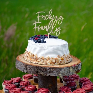 Op de afbeelding: Een wit geglazuurde taart met blauwe bessen en frambozen, met een houten topper met de tekst "Fucking Finally". De taart staat op een houten standaard, omringd door cupcakes met rode glazuur.