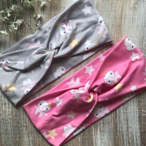 May include: Two fabric headbands with a twisted knot design. One headband is gray with white stars and a white tooth with a crown design. The other headband is pink with white stars and a white tooth with a crown design.