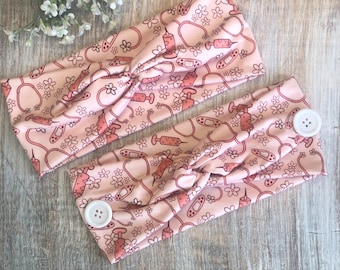 May include: Two pink fabric headbands with a white button on each end. The headbands have a repeating pattern of pink flowers, bandages, and syringes.
