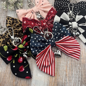 Pode incluir: Uma coleção de chaveiros com laços de tecido em vários padrões, incluindo bolinhas, cerejas, estampa de leopardo e um design de bandeira americana. Cada laço é preso a um fecho prateado e um pingente em forma de coração. Dispostos sobre uma superfície de madeira clara.