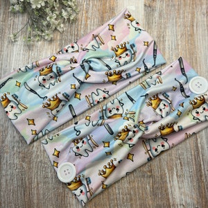 May include: Two colorful headbands with a tooth design. The headbands have a white button on one end and a twist design in the middle. The tooth design features a tooth with a crown and a toothbrush.