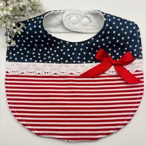 May include: A baby bib with a patriotic design. The top portion is navy blue with white stars, the middle has white lace, and the bottom is red and white striped. A red bow is attached. The bib has a white terry cloth backing and two white snap closures.