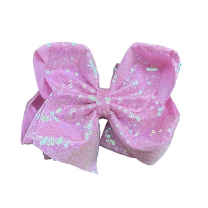 May include: A large, light pink hair bow with a sequined finish. The bow is made of a soft fabric and features a classic bow shape. The sequins are iridescent, adding a subtle shimmer to the accessory.
