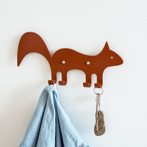 May include: A brown metal squirrel-shaped wall hook with three hooks. A light blue shirt hangs from one hook, and a set of keys with a flip-flop keychain hangs from another. The hook is mounted on a white wall.