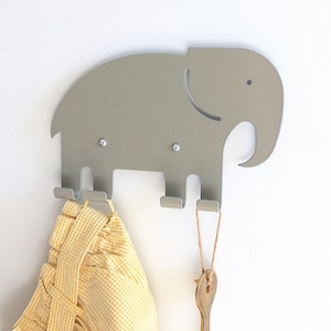May include: A grey metal elephant-shaped wall hook with four hooks. A yellow and white checked fabric is hanging on one of the hooks. A wooden spoon is hanging on another hook.