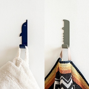 Pode incluir: Dois ganchos de parede, um com a forma de um golfinho em azul escuro e o outro com a forma de uma serra em verde claro, com toalhas penduradas.