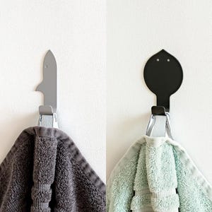 May include: Two wall hooks, one shaped like a fish and one shaped like a leaf, with towels hanging on them. The fish hook is gray and the leaf hook is black.