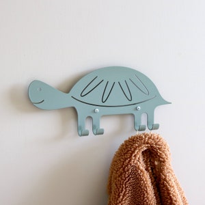 May include: A light blue metal turtle-shaped wall hook with three hooks. A brown fuzzy towel is hanging on one of the hooks.