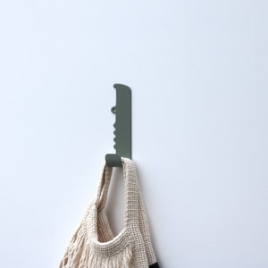A bathroom wall hook with the shape of a crocodile, seen from the side and facing right. It is made out of stainless steel and painted with a green color. The crocodile´s body has been bent up in to one hook, to put backpacks, jackets and keys on.