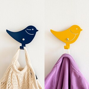 May include: Two metal bird-shaped hooks on a white wall. One hook is blue and the other is yellow. A white mesh bag hangs from the blue hook and a purple shirt hangs from the yellow hook.