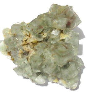 May include: A cluster of pale green fluorite crystals with a translucent appearance. The crystals have a cubic shape and are arranged in a layered formation. The base of the cluster has a yellowish-brown matrix.