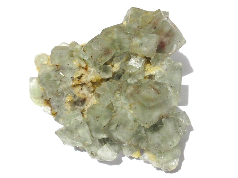 May include: A cluster of pale green fluorite crystals with a translucent appearance. The crystals have a cubic shape and are arranged in a layered formation. The base of the cluster has a yellowish-brown matrix.