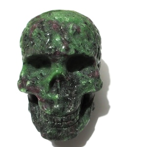 May include: A carved skull made of green and maroon stone. The skull has detailed features, including eye sockets, a nose cavity, and teeth. The stone has a rough texture and a natural, organic appearance. The skull is set against a white background.