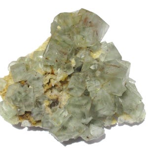 May include: A cluster of pale green fluorite crystals with a cubic structure. The crystals are translucent, with hints of red and brown inclusions. The fluorite sits on a matrix of beige and yellow minerals, creating a natural mineral specimen.